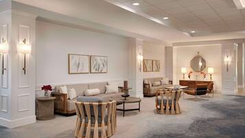 A spacious hotel lobby with a neutral color palette, featuring a seating area with sofas and chairs, a coffee table, and a decorative mirror.