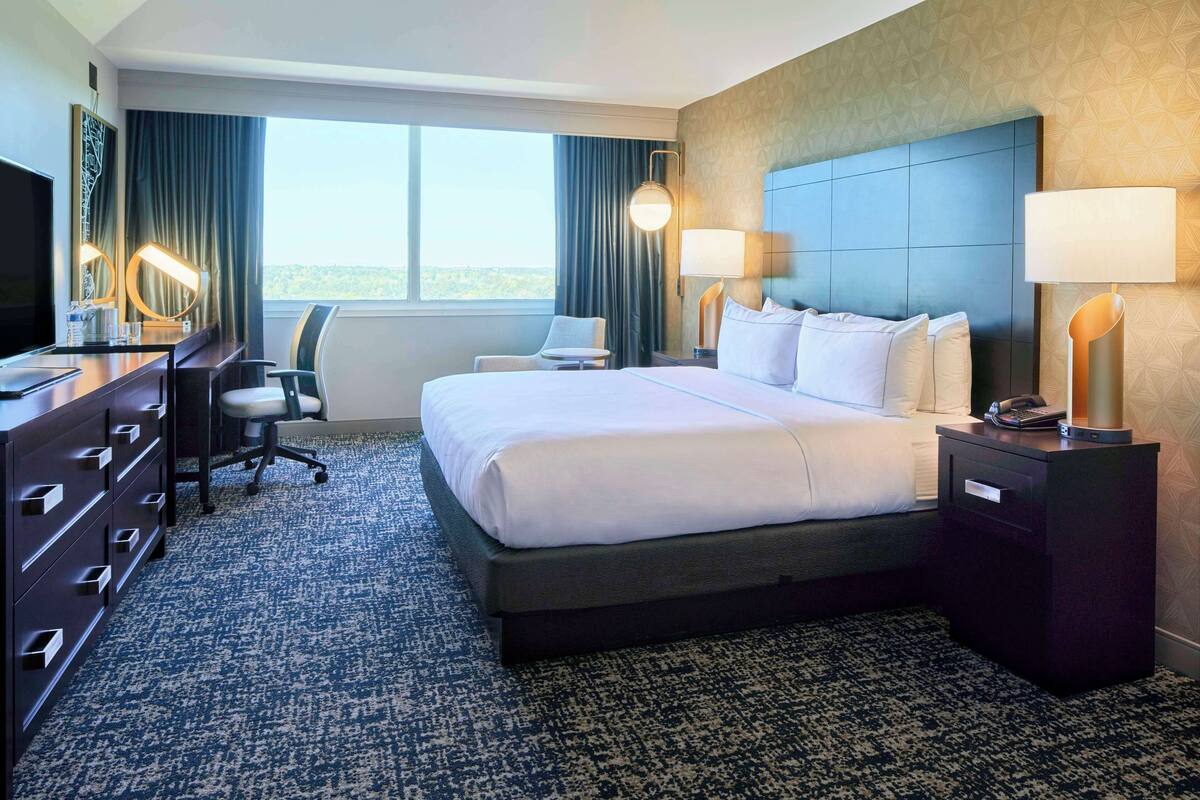 A hotel room with a large bed, a desk with a computer, a chair, a TV, and a window with a view.