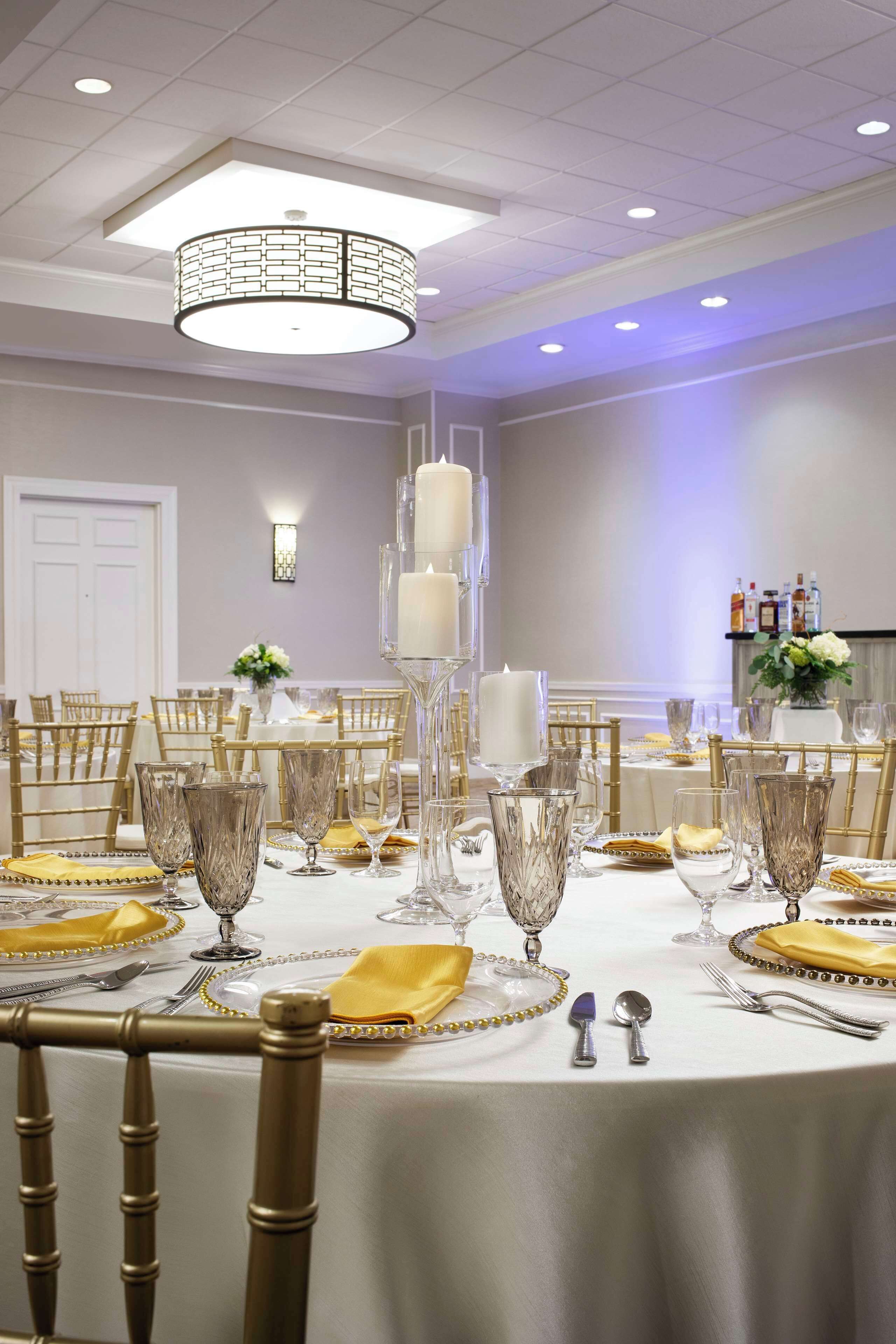 Un salón de banquetes con mesas redondas dispuestas para comer, con sillas doradas, manteles blancos y cristalería de cristal.
