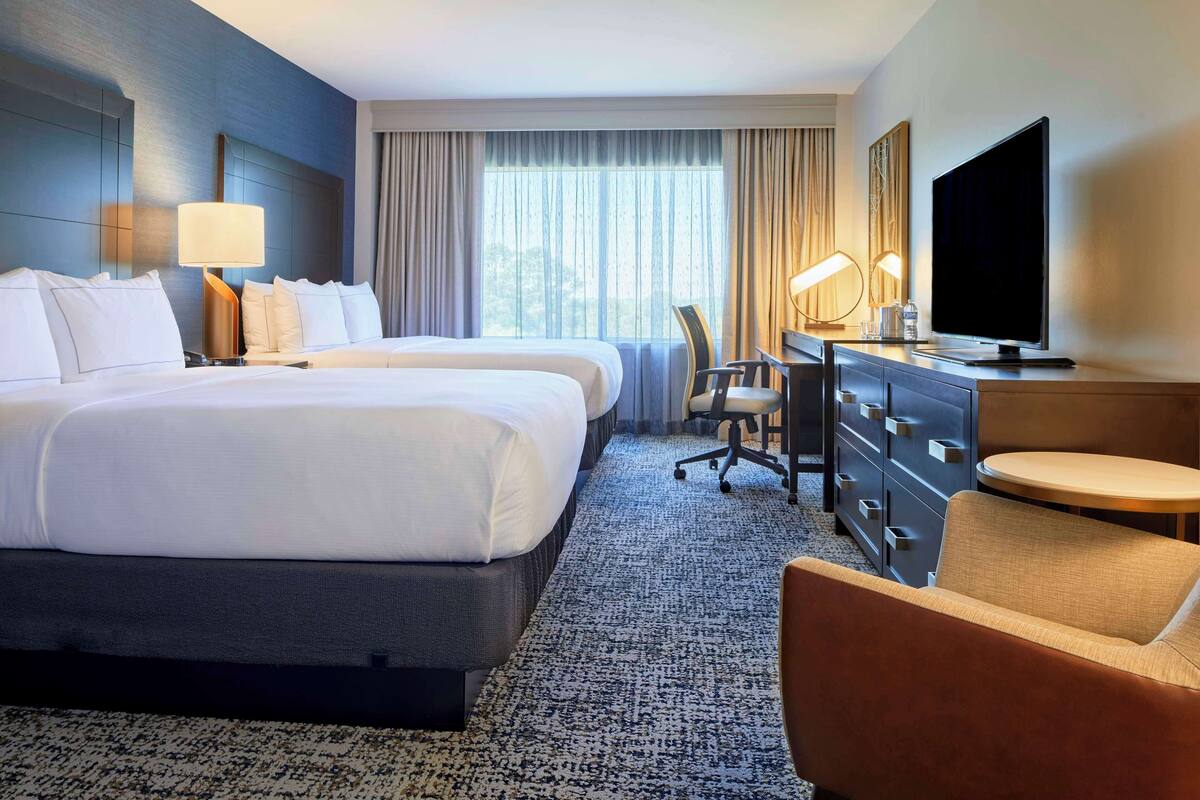A hotel room with two beds, a desk with a chair, a TV, and a small round table.