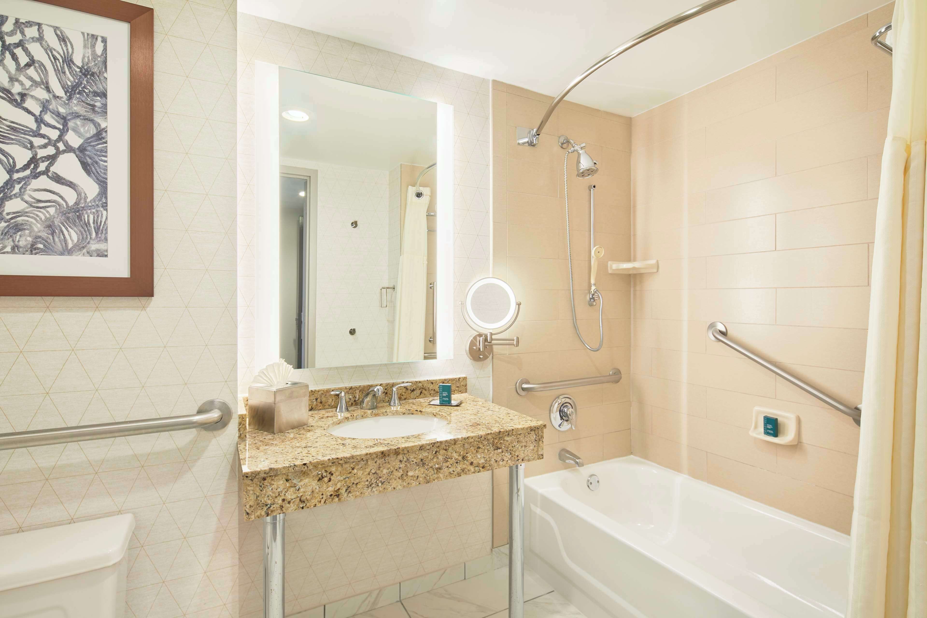 A modern bathroom with a granite countertop sink, a large mirror, a handheld showerhead, a handheld shower, a bathtub, and a toilet.
