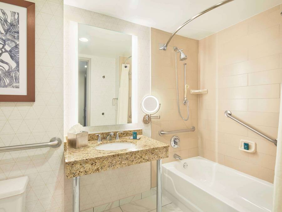 A modern bathroom with a granite countertop sink, a large mirror, a handheld showerhead, a handheld shower, a bathtub, and a toilet.