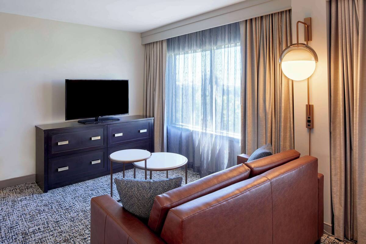 A hotel room with a leather sofa, a flat-screen TV, a small table, and a lamp.