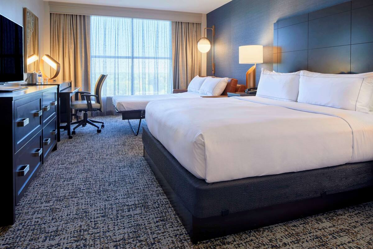 A hotel room with a large bed, a desk with a chair, a TV, and a window with curtains.