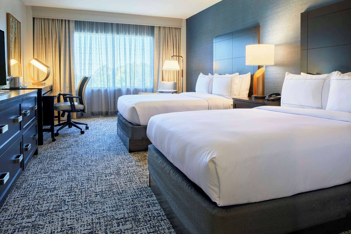 A hotel room with two beds, a desk, a chair, and a large window with curtains.