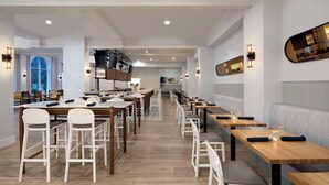 A modern restaurant with multiple dining tables and chairs, a long counter, and a view of the outdoors through a large window. - DoubleTree by Hilton Atlanta - Roswell (Roswell)