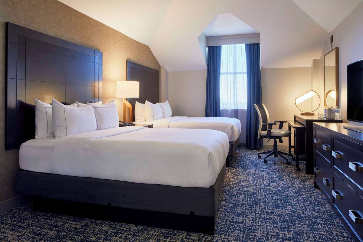 A hotel room with two beds, a desk with a chair, a dresser, a TV, and a large window with curtains.