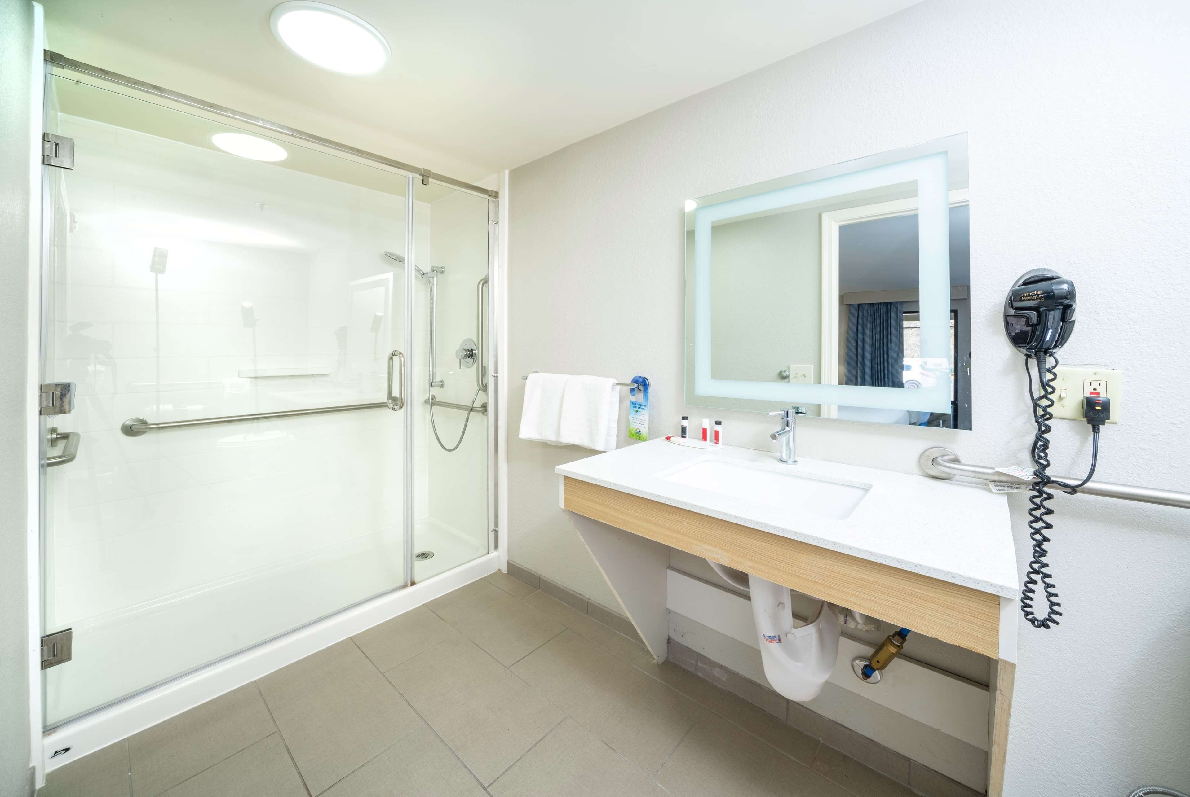Un baño moderno con cabina de ducha de vidrio, un lavamanos blanco con encimera de madera y un espejo con teléfono integrado.