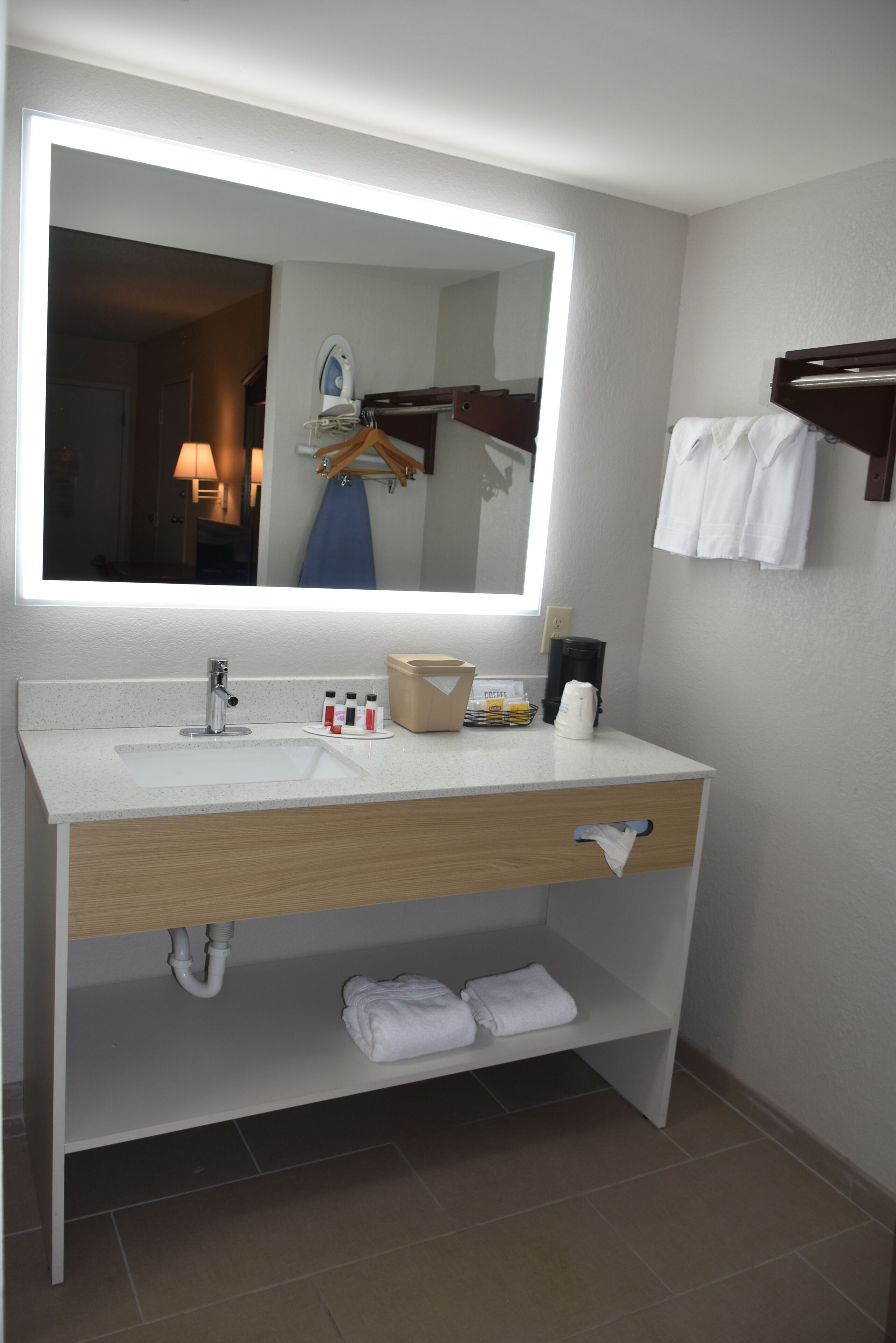 A modern bathroom with a large mirror, a white sink, a towel rack, and a shelf with toiletries.