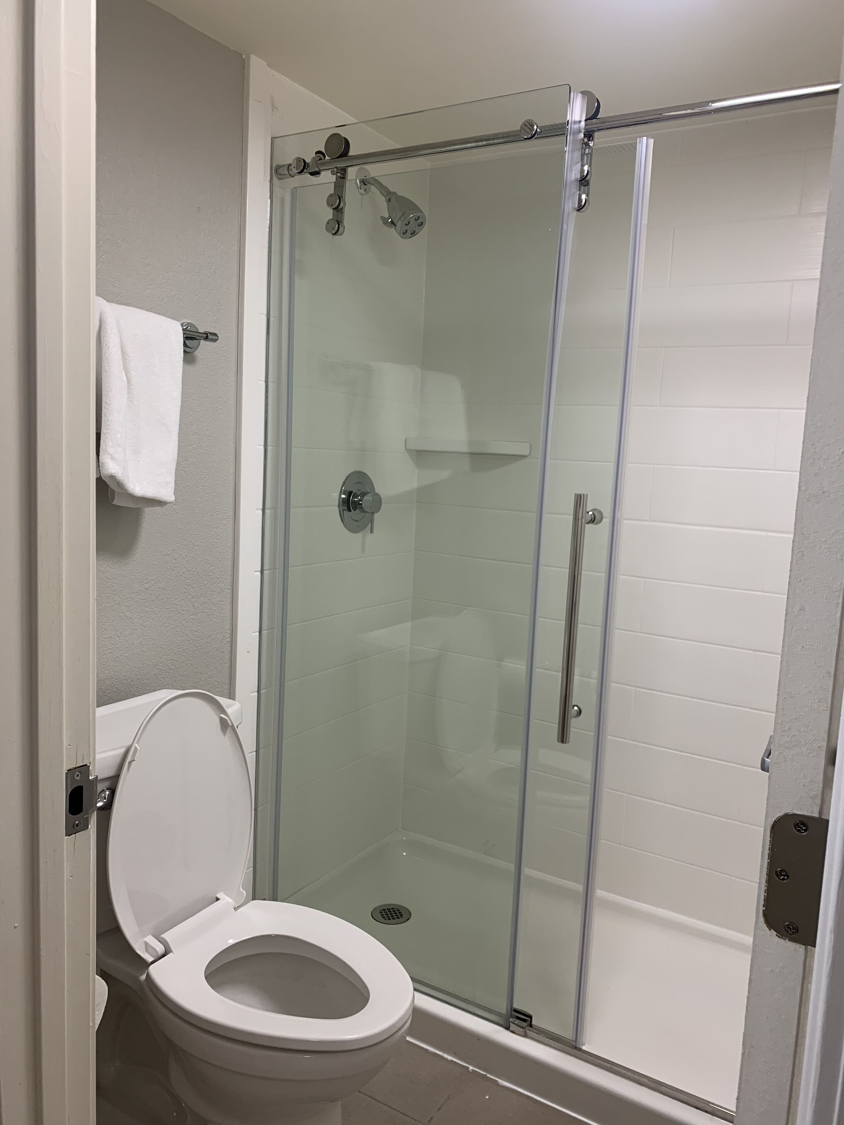 A glass-enclosed shower stall with a white tiled wall, a white toilet with a closed lid, and a white towel hanging on a rack.