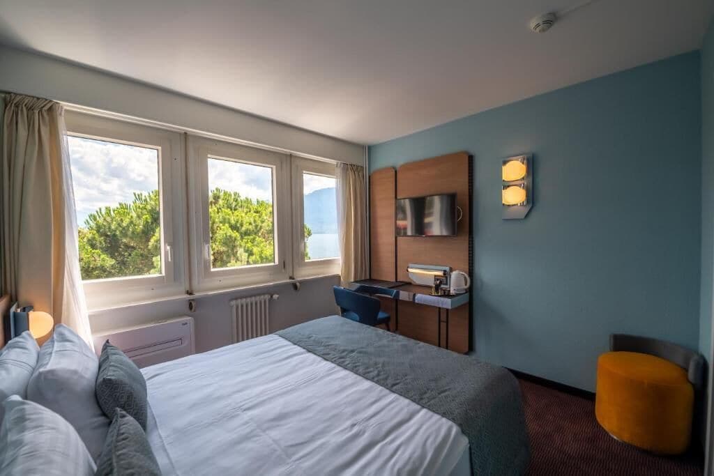 A hotel room with a bed, a desk, a chair, a TV, and a window with a view of trees.
