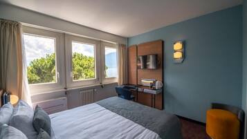 A hotel room with a bed, a desk, a chair, a TV, and a window with a view of trees.