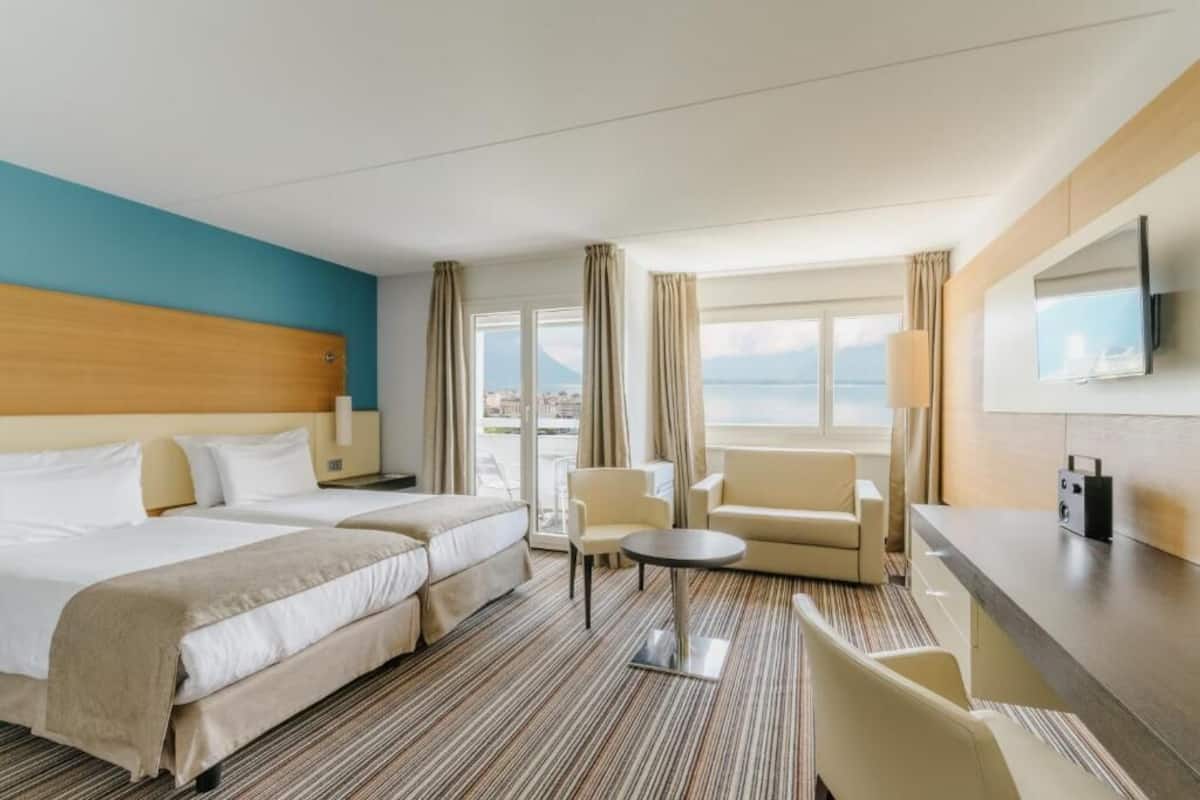 Habitación de hotel con dos camas, un sofá, una mesita, televisión y vista al mar.