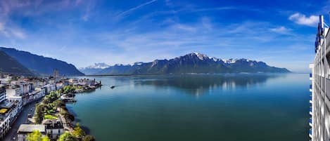 A coastal city with a calm lake, mountains in the background, and a modern building with balconies on the right.