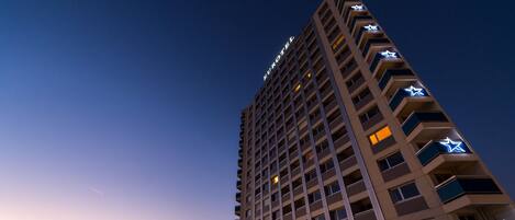 Un hotel alto de diseño moderno, con balcones con luces en forma de estrella y vistas al atardecer sobre un cuerpo de agua.