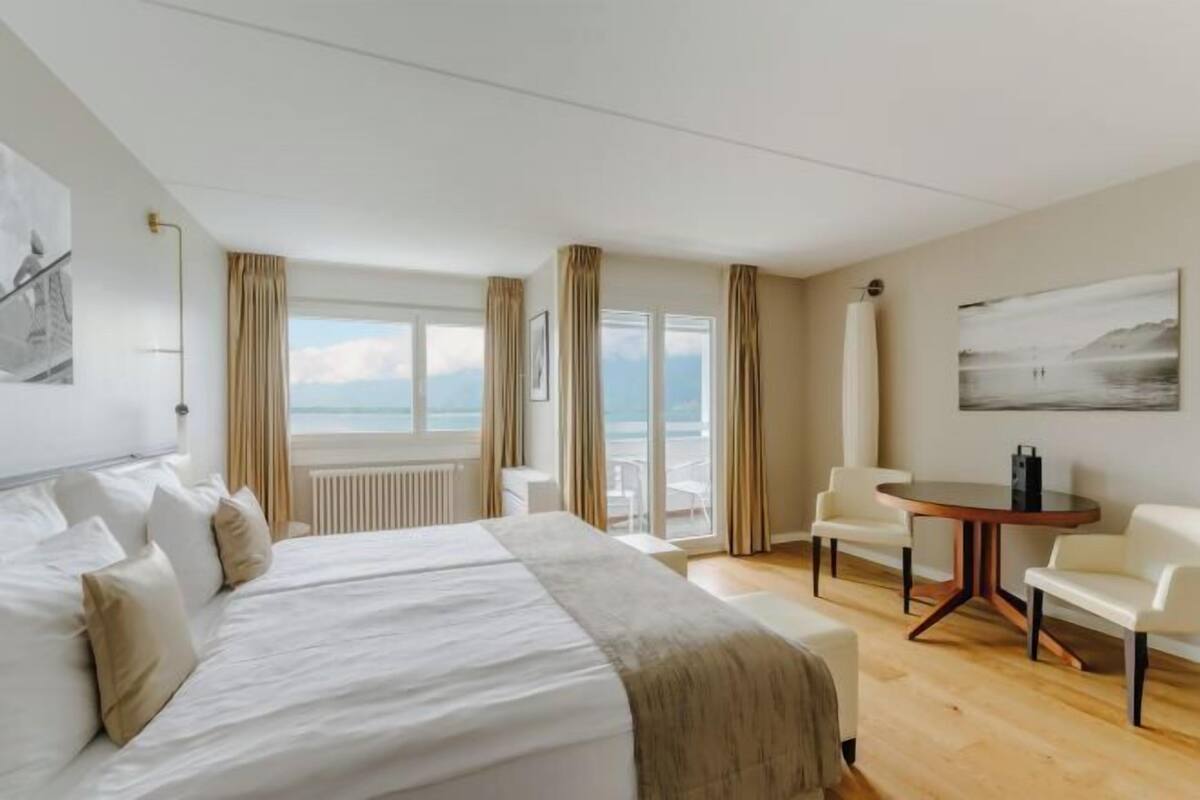 A hotel room with a bed, a round table, two chairs, a view of the sea, and a painting on the wall.