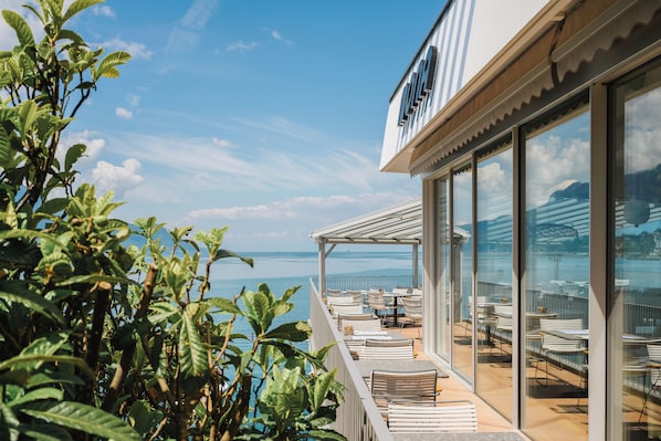 A balcony with outdoor seating overlooking a body of water, a clear sky, and a view of a bridge in the distance. - Mona Montreux (Montreux)