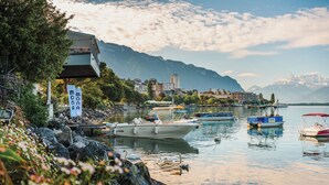 A lakeside view with boats docked, a building with a balcony, and mountains in the background.