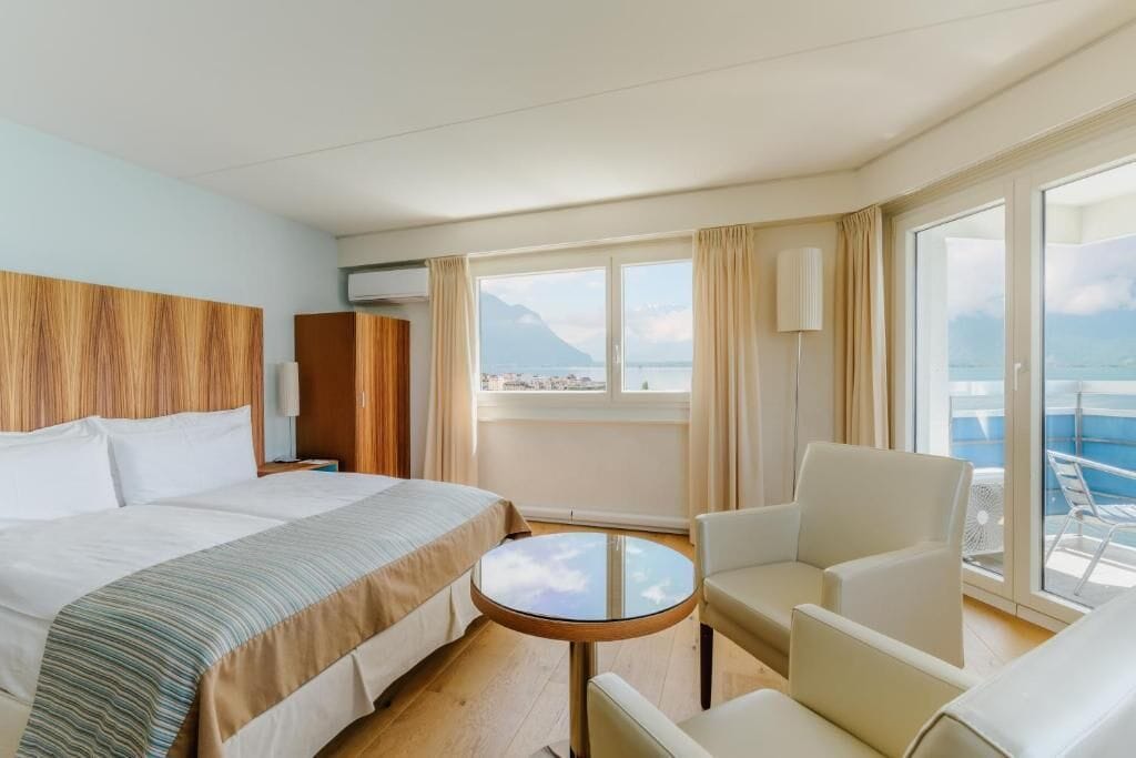 A hotel room with a bed, a small table, two chairs, a window with a view of the sea, and a balcony.
