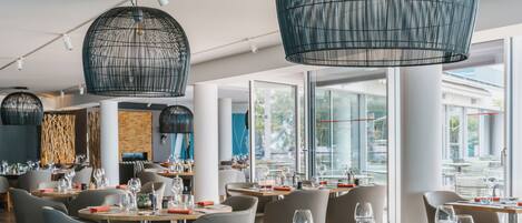 A modern restaurant with round tables, grey chairs, and black hanging light fixtures.