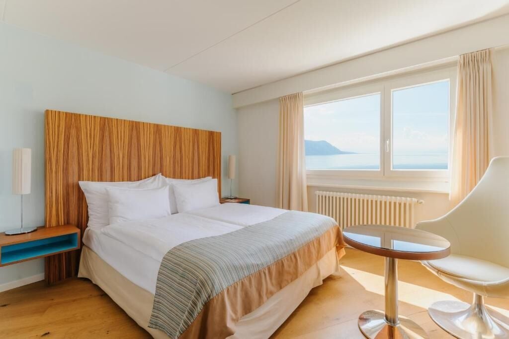 A hotel room with a bed, a round table, a chair, a nightstand, and a view of the sea.
