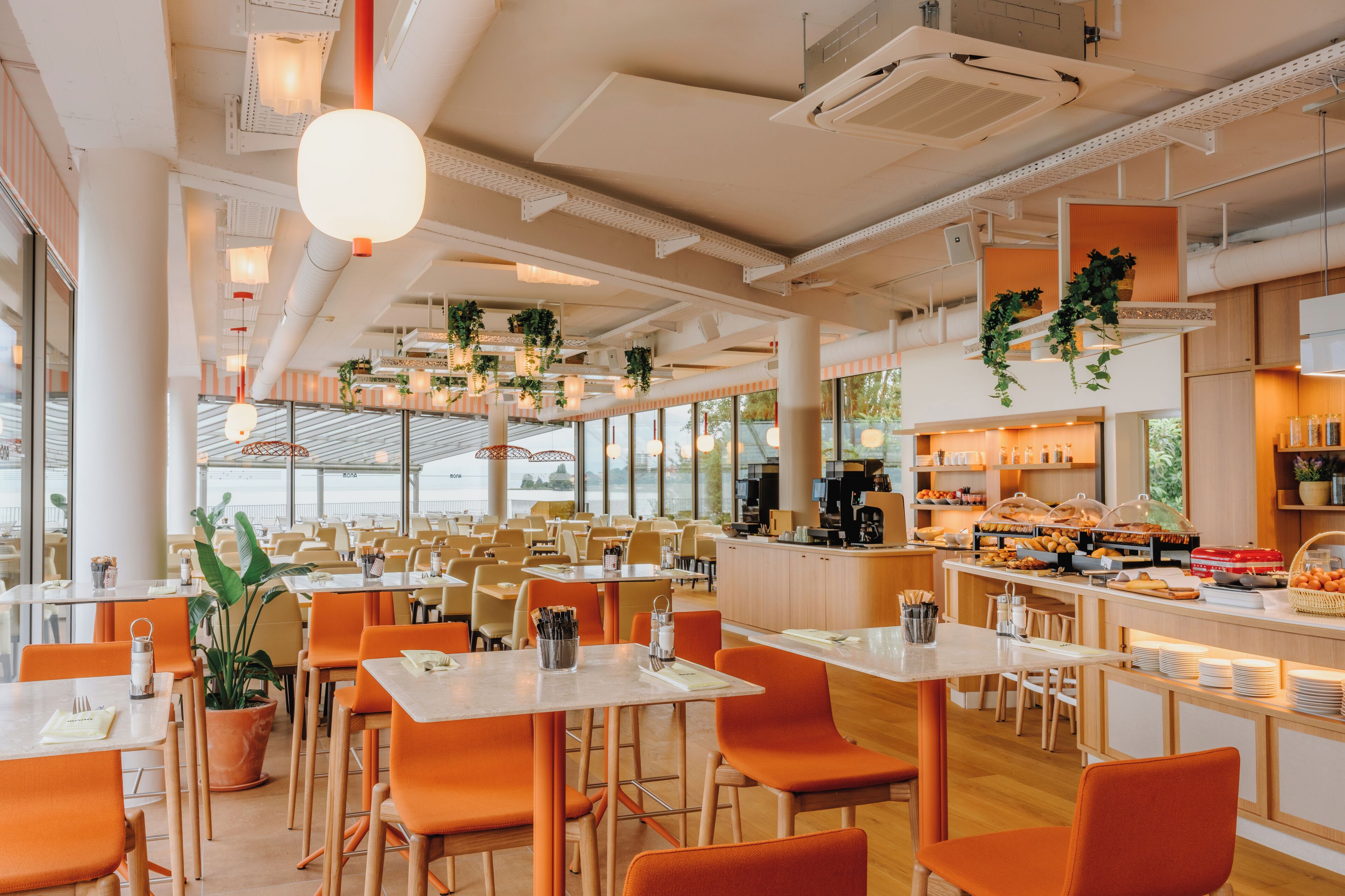 Un comedor amplio con varias mesas y sillas, una barra de buffet con alimentos, y ventanas grandes que permiten la entrada de luz natural.