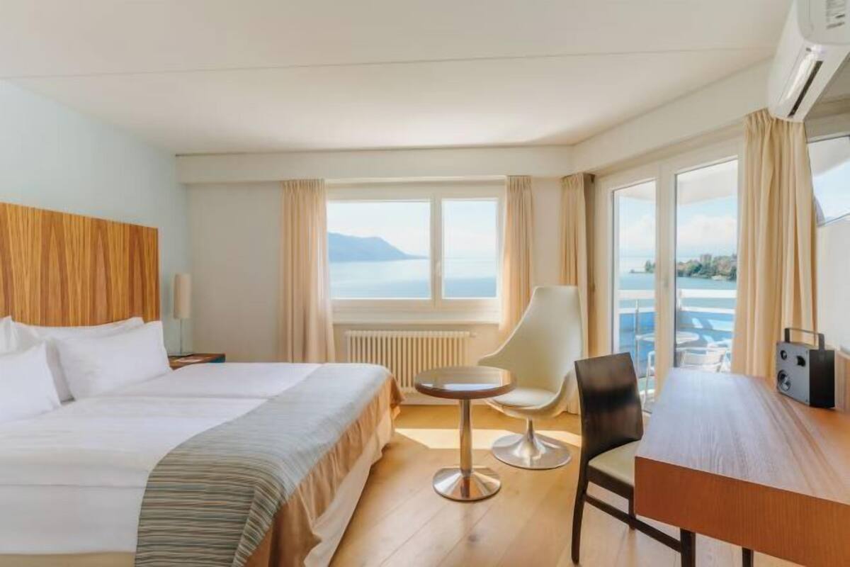 A hotel room with a bed, a round table, a chair, a wooden desk, and a view of the sea.