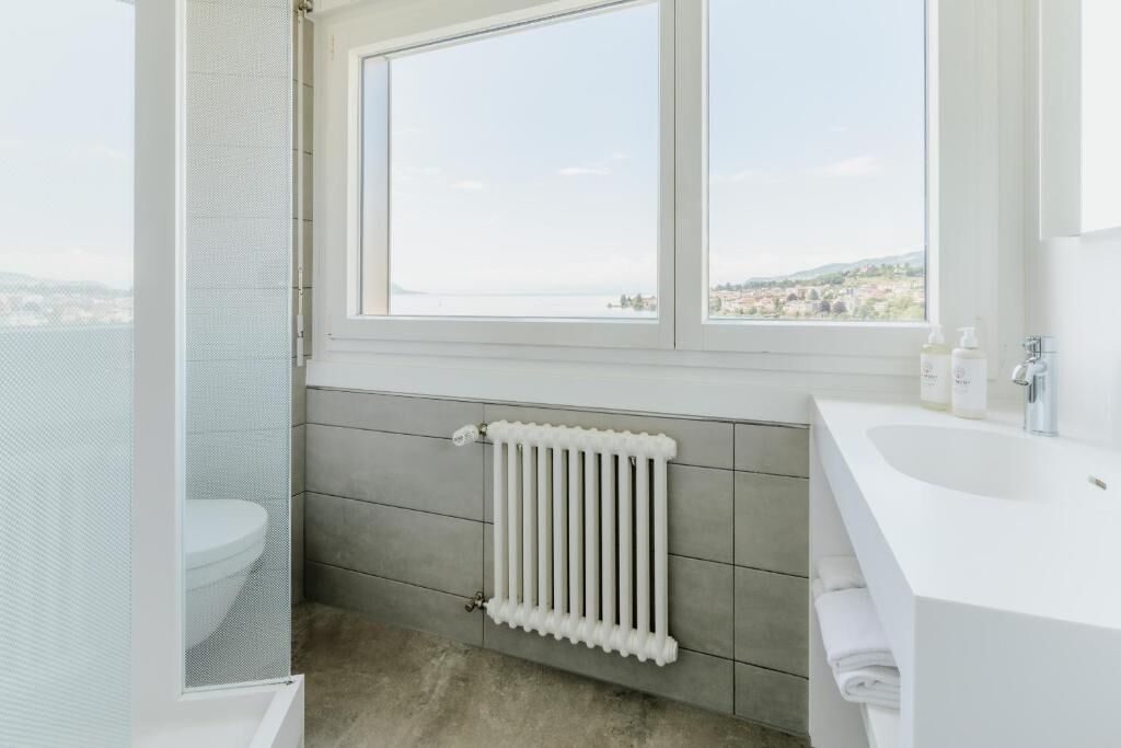 Une salle de bain avec des toilettes, une fenêtre donnant sur la ville, un radiateur blanc et un lavabo avec des articles de toilette.