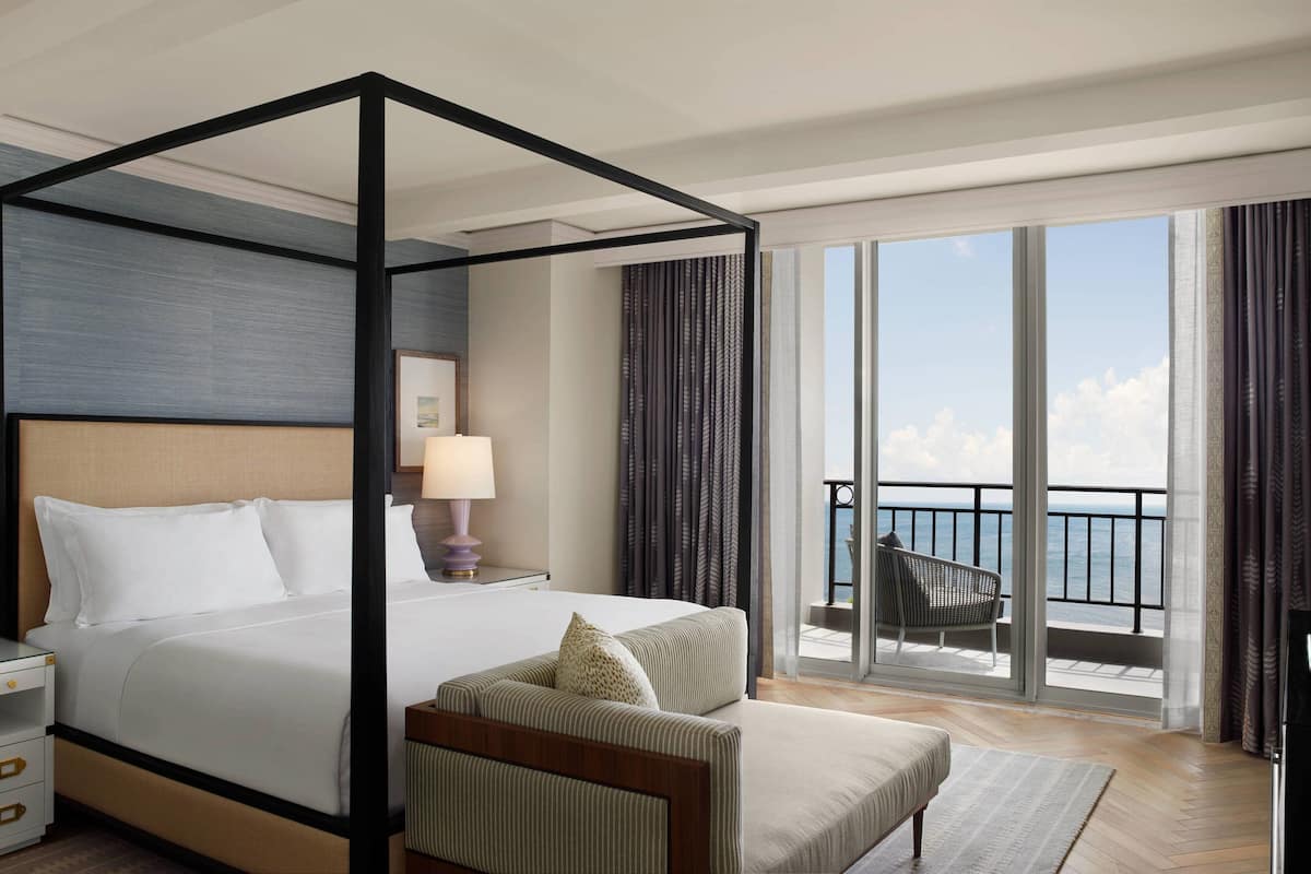 A four-poster bed with a view of the ocean, a balcony with a chair, and a nightstand with a lamp.