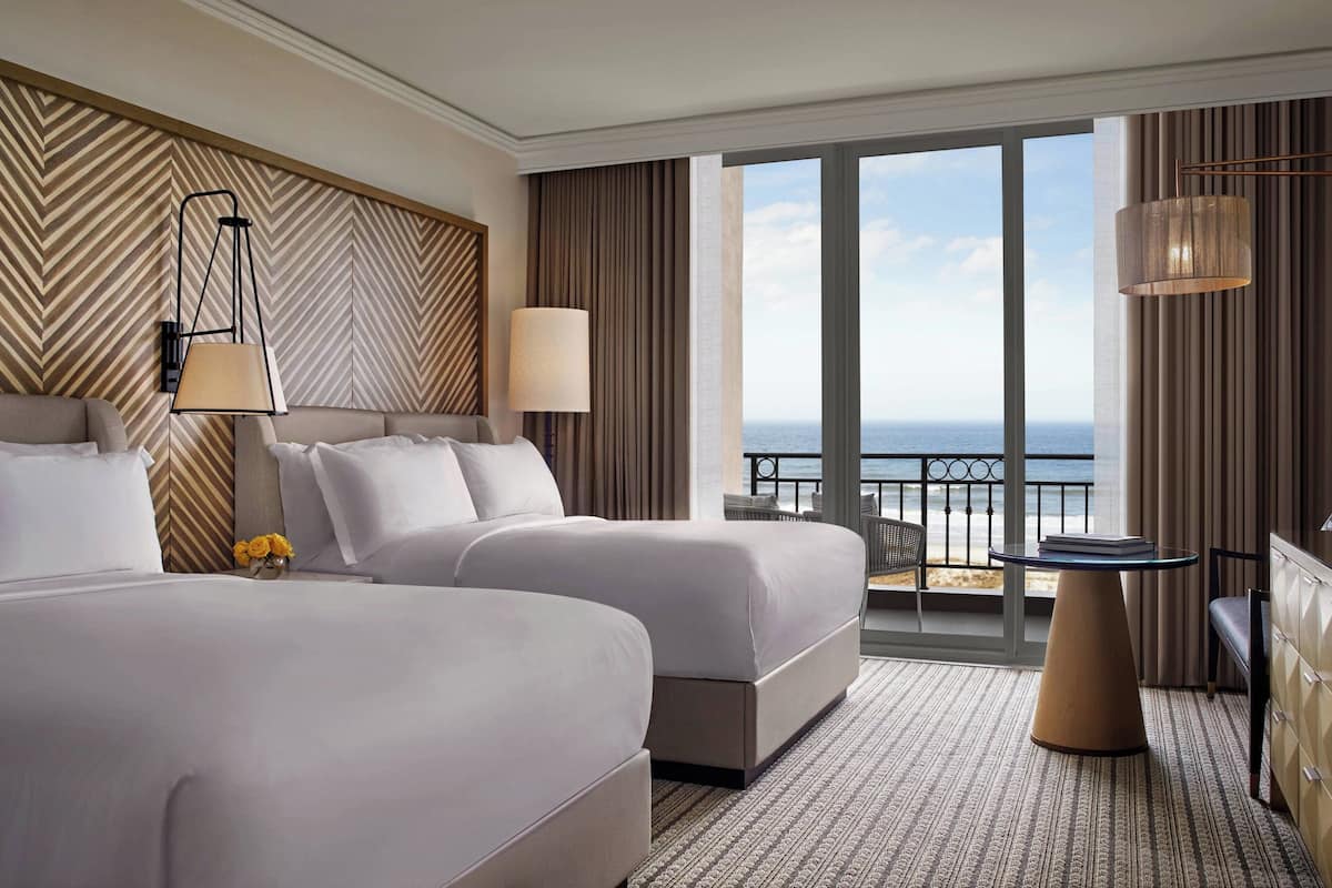 A hotel room with two beds, a balcony with a view of the sea, and a table with a lamp.