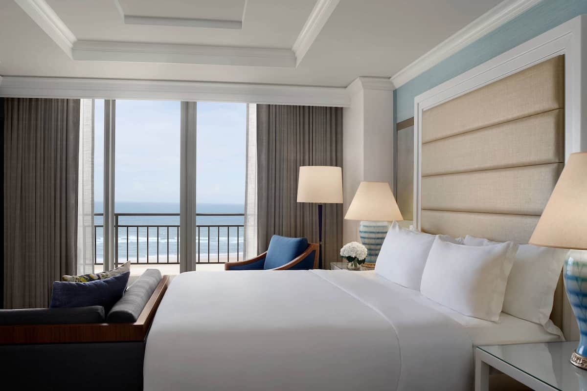 A hotel room with a large bed, two bedside lamps, a chair, and a view of the ocean.