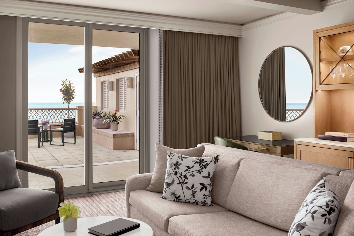 A modern hotel room with a balcony, a sofa, a coffee table, and a desk with a mirror and a wooden cabinet.