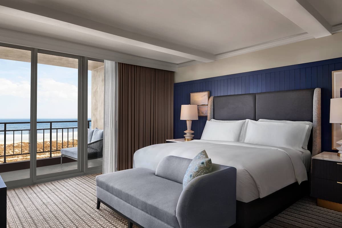 A hotel room with a large bed, a desk, a chair, a nightstand, and a view of the beach.