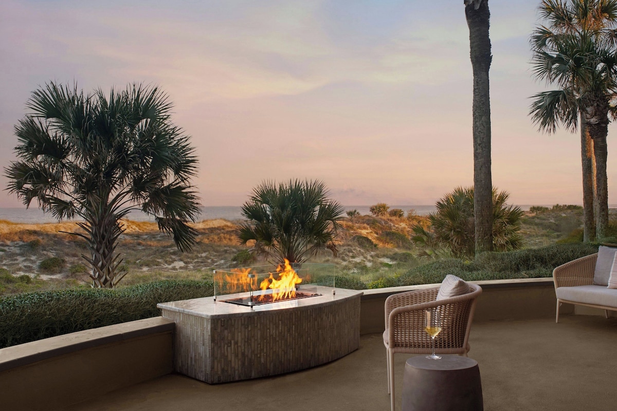 A fire pit with a clear glass enclosure, surrounded by wicker furniture and palm trees, overlooking a beach.