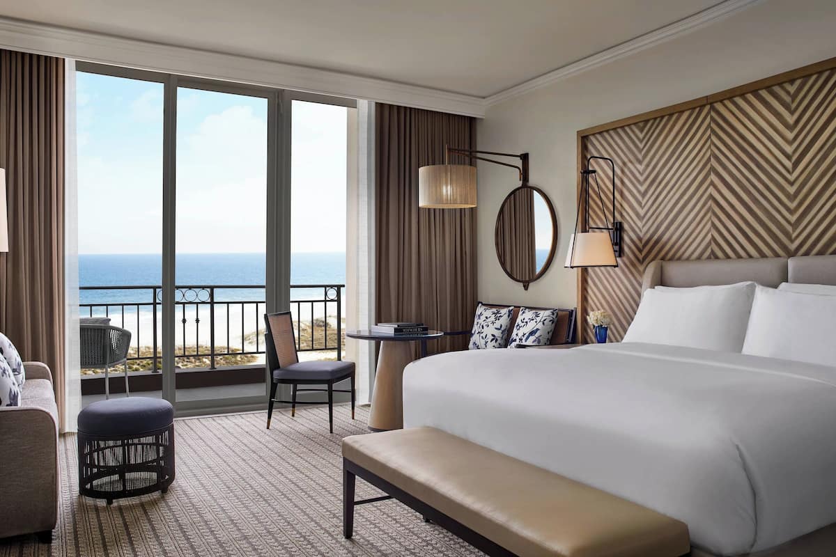 A hotel room with a large bed, a bench, a chair, a small table, and a view of the ocean through large windows.