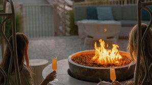 Two people sitting on a swing chair with a fire pit table between them, overlooking a sunset.