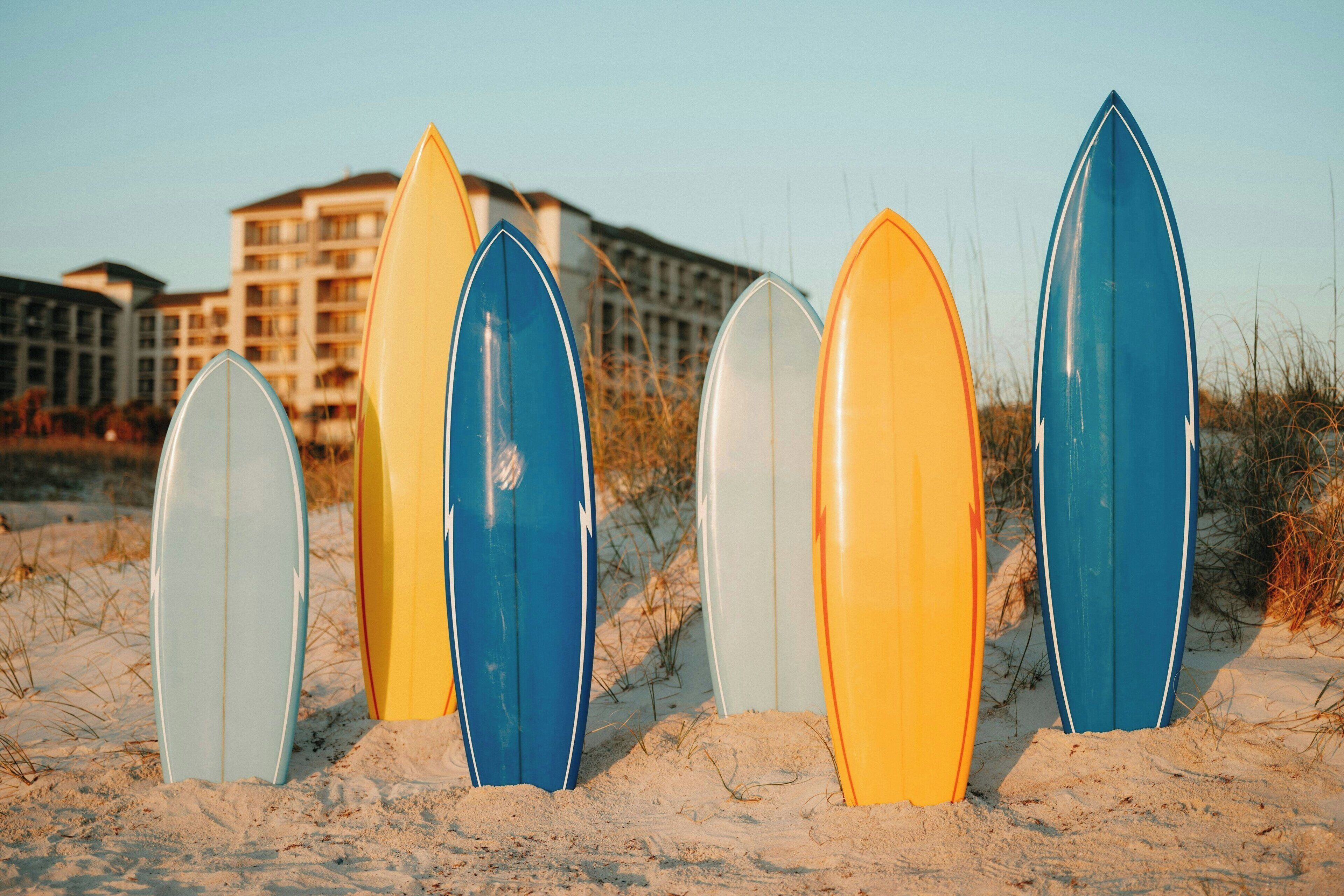 Eine Reihe von Surfbrettern an einem sandigen Strand, im Hintergrund Gebäude.
