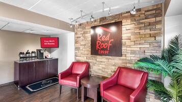 A Red Roof Inn lobby with a stone wall, two red leather chairs, a coffee station, and a flat-screen TV displaying 'Free Wi-Fi All Rooms'.