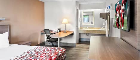 Habitació d'hotel amb llit, escriptori, cadira, làmpada, televisió i un bany amb mirall i benedora.