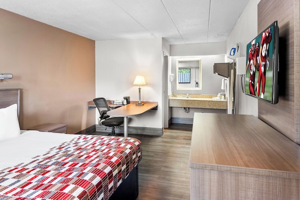 A hotel room with a bed, desk, chair, lamp, television, and a bathroom with a mirror and sink.