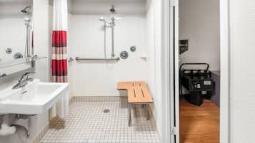 A bathroom with a white sink, a shower with a red and white curtain, a wooden bench, and a black luggage trolley.