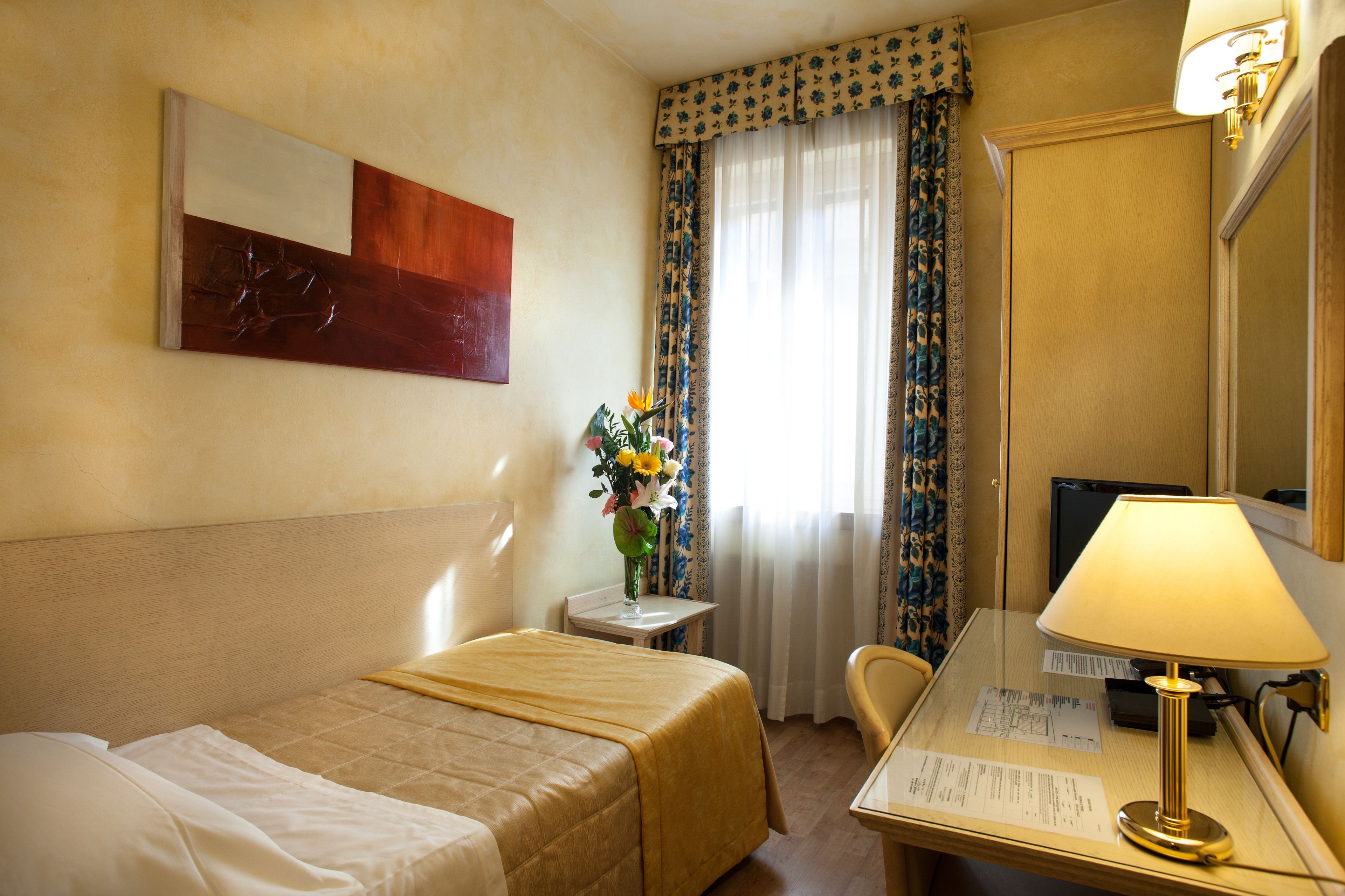 Habitación de hotel con dos camas, un escritorio, una lámpara, un florero con flores y una ventana con cortinas.