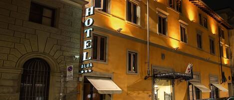 Un hotel llamado "HOTEL ALBA" de estilo arquitectónico clásico, con una entrada destacada, un estacionamiento visible y un letrero que indica zona de no estacionamiento.