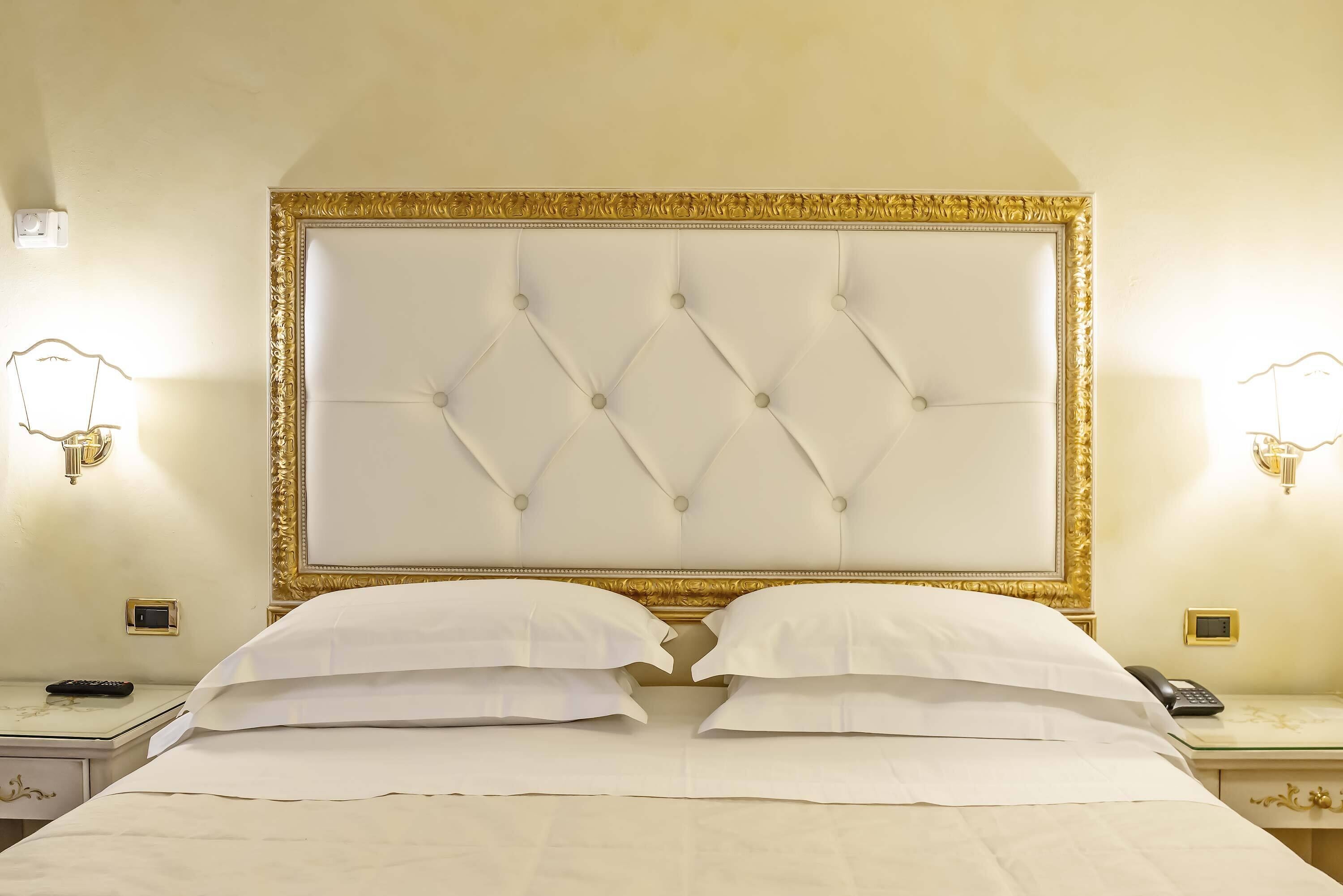 Una habitación de hotel con un cabecero capitonado, dos mesitas de noche con lámparas, y una cama con ropa blanca.