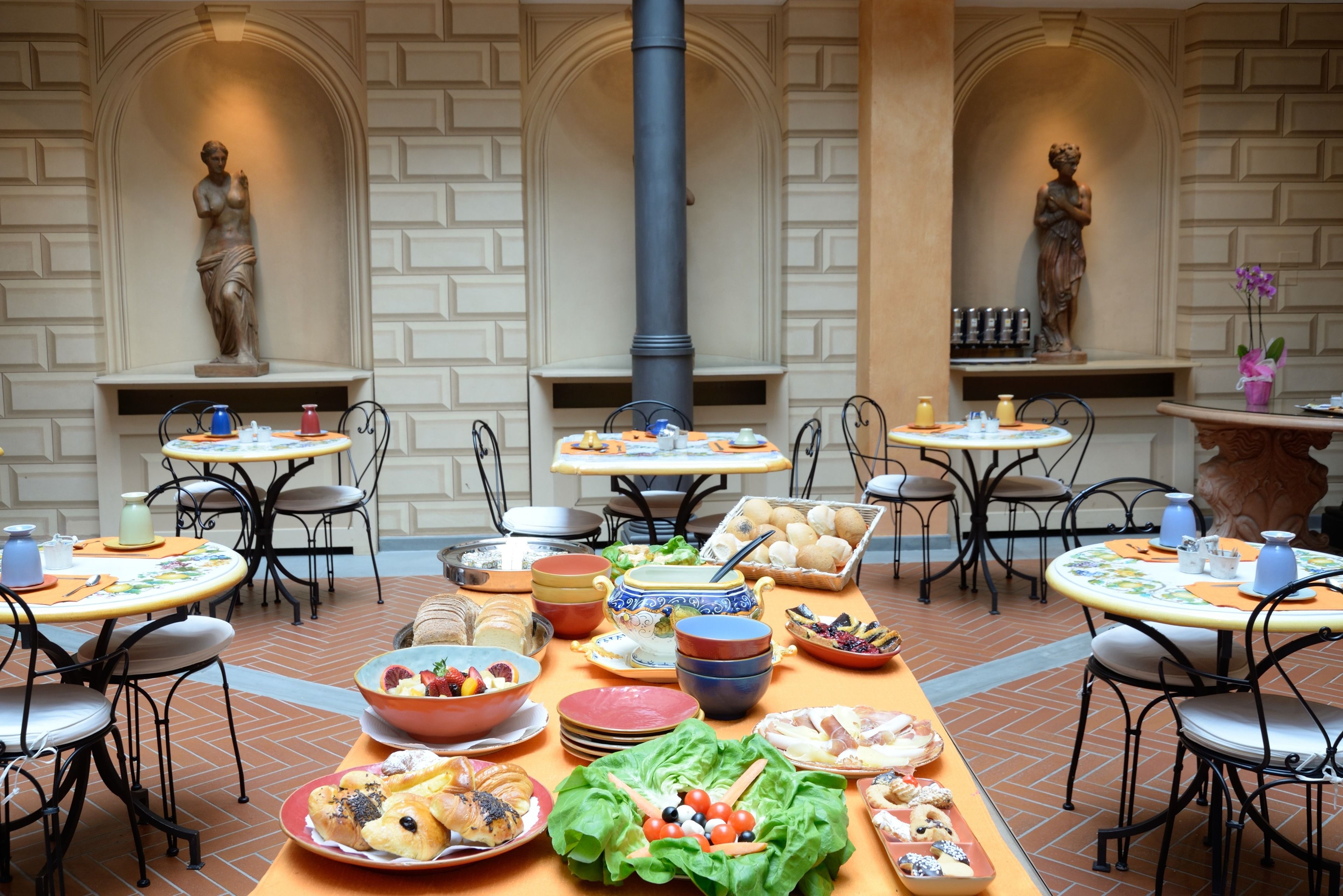A dining area with multiple tables and chairs, a variety of food and drink items, and decorative statues.