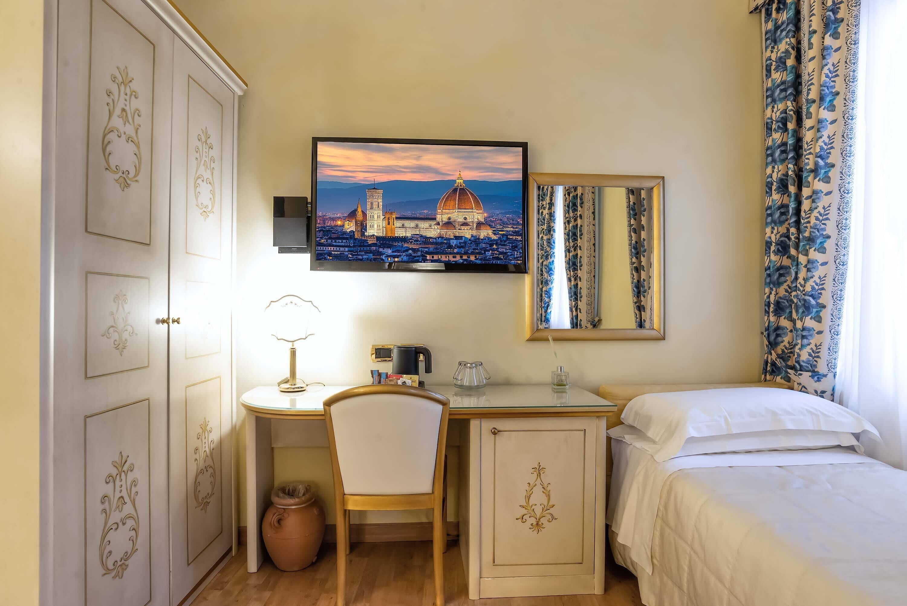 Un dormitorio con cama, un escritorio con silla, un televisor y un amplio ventanal con cortinas.