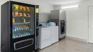 Ein Raum mit Getränke- und Snackautomaten, einer Waschmaschine und einem Trockner, außerdem ein Fliesenboden und eine weiße Tür.