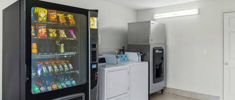 Une machine distributrice de collations et de boissons, un lave-linge et un sèche-linge se trouvent dans une pièce dont le sol est carrelé et la porte est blanche.