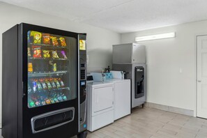 Une machine distributrice de collations et de boissons, un lave-linge et un sèche-linge se trouvent dans une pièce dont le sol est carrelé et la porte est blanche.