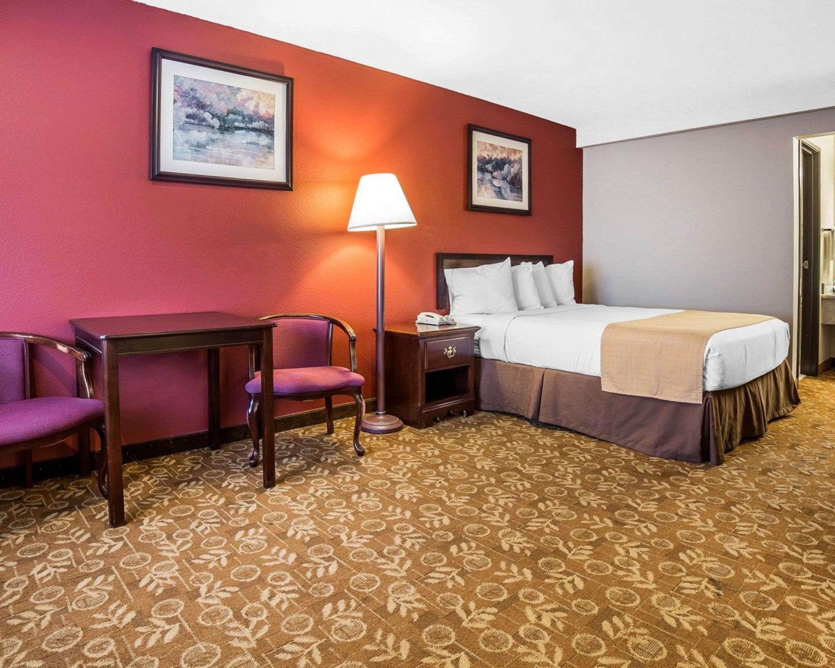 A hotel room with a bed, a desk, a chair, a lamp, and two framed pictures on the wall.